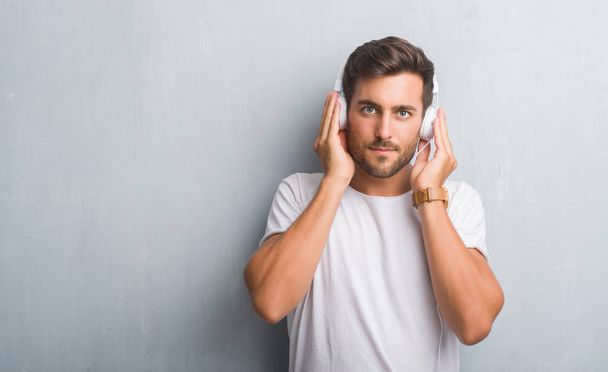 Pohledný mladý muž přes zeď šedá grunge sluchátka poslouchat hudbu s výrazem věří na inteligentní tvář myslí vážně - Fotografie, Obrázek