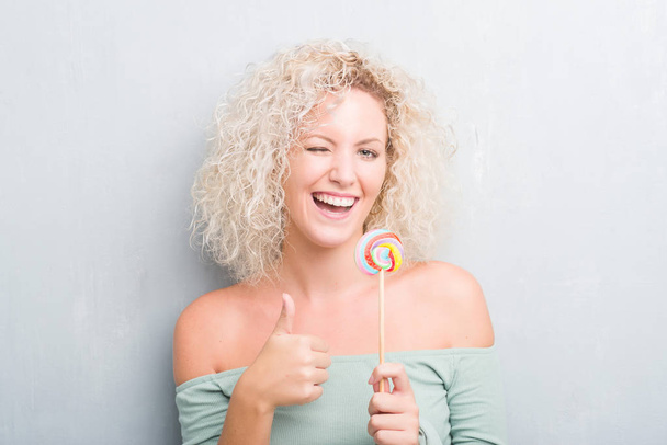 Νεαρή ξανθιά γυναίκα πάνω από το grunge γκρίζο τοίχο τρώει candy lollipop ευτυχισμένος με μεγάλο χαμόγελο κάνει εντάξει σήμα, τον αντίχειρα επάνω με τα δάχτυλα, καλό σημάδι - Φωτογραφία, εικόνα