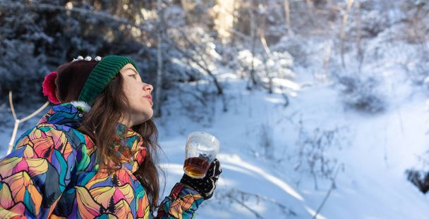 Girl enjoys the snow falls. Young woman in a knitted shape is drinking tea in the forest during a snowfall. Toned photo - Photo, Image