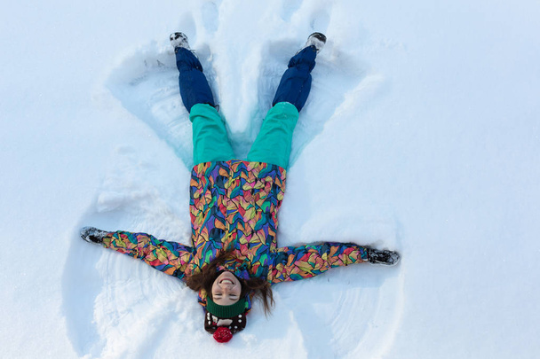 Υψηλή γωνία άποψη του ευτυχισμένου κοριτσιού που βρίσκεται στο χιόνι και κινείται τα χέρια και τα πόδια της πάνω και κάτω δημιουργώντας μια φιγούρα άγγελος χιόνι. Χαμογελαστή γυναίκα ξαπλωμένη στο χιόνι στις χειμερινές διακοπές με αντίγραφο χώρου - Φωτογραφία, εικόνα