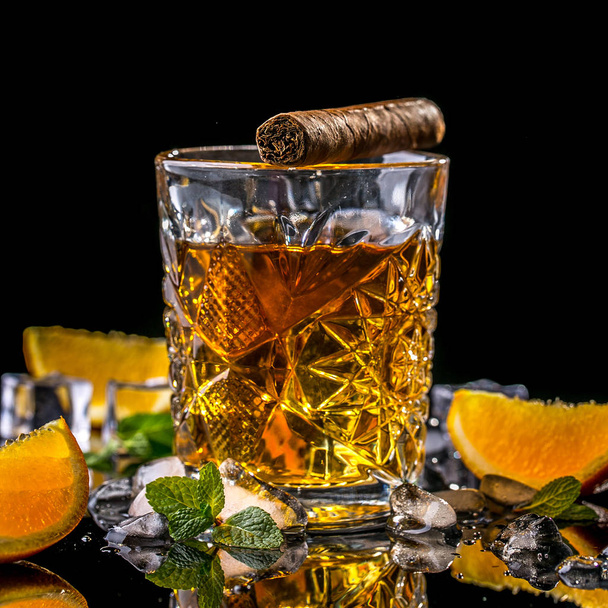 Stillleben-Komposition mit Glas Whiskey und Zigarre mit Eis und Minze auf schwarzem Hintergrund. Konzept Luxus-Drink-Cocktail. - Foto, Bild
