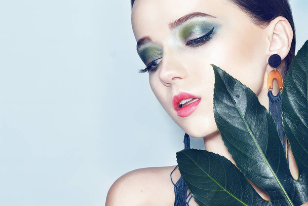 Sexy beauty brunette model with green and blue make up and green leaves. Copy space. Close up, selective focus. Girl with tropical leaf covers a part of her face. Fashion, cosmetics, accessories. - Photo, Image