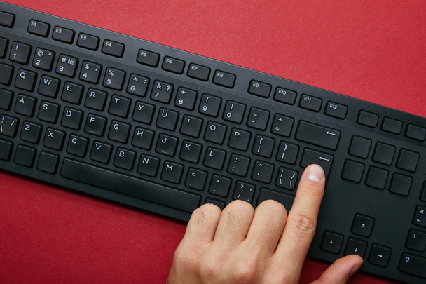 Ansicht von oben: Mann drückt Taste auf schwarzer Computertastatur auf rotem Hintergrund  - Foto, Bild