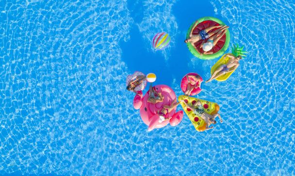 AERIAL TOP DOWN COPY SPACE: підходить дівчатам і м'язистим хлопцям, які лежать на барвистих бочках у басейні, грають у волейбол з м'ячем. Друзі на веселих літніх канікулах плавають на воді на надувних шезлонгах
 - Фото, зображення