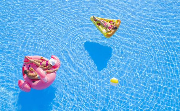 AERIAL CLOSE UP Красива пара і підійде дівчині, що лежить на веселому барвистому надувному плаву, насолоджуючись спокійною літньою відпусткою на курорті. Друзі охолоджують, посипають напої та сонячні ванни на плаву в басейні
 - Фото, зображення