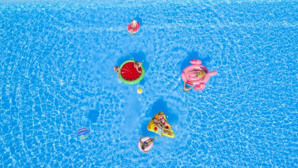 AERIAL: Cheerful happy friends having water gun fight on colorful floaties at summer pool party. Playful guys and girls splashing water on inflatable pizza, doughnut, flamingo and watermelon floats - Photo, Image