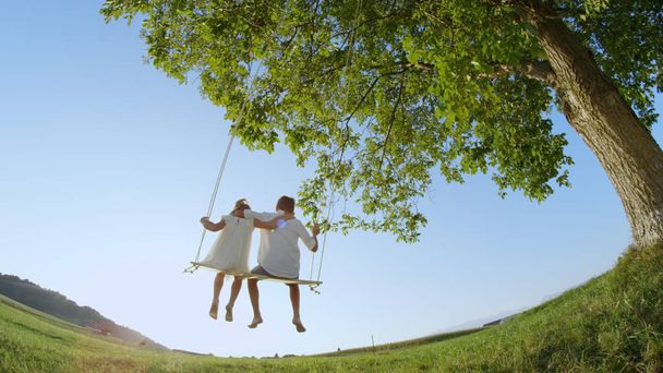 LOW ANGLE VIEW: Couple of young kids holding each other swinging on a wooden swing under a big green tree in summer. Boy and girl in embrace on a warm day. Hugging siblings swaying barefoot. - Foto, immagini