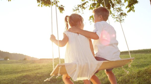 CLOSE UP LENS FLARE Couple children embracing on wooden swing at gold sunset. Young love blooming between boy and girl swinging on swing set. Brother hugging his little sister enjoying a warm evening - Фото, зображення