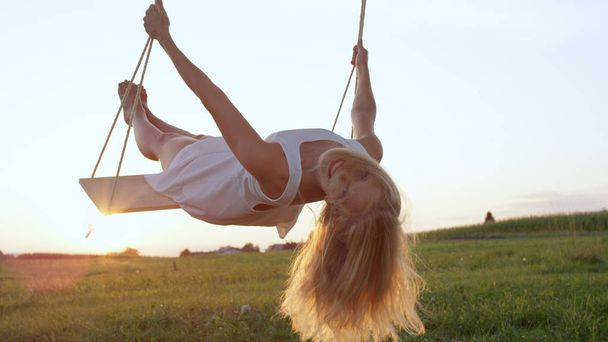 LENS FLARE CLOSE UP: Happy young blonde woman in white dress looking back and smiling into the camera while swinging in golden summer sunset in country meadow. Cheerful lady leaning back in her swing - Foto, Bild