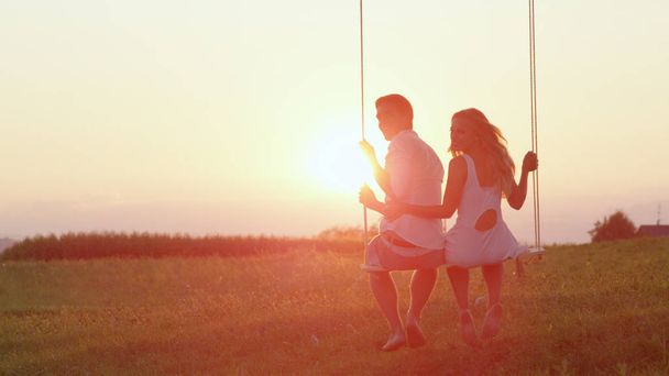 Linsenschlag: entzücktes lächelndes Paar, das sich während eines romantischen Sonnenuntergangs auf einer Schaukel wiegt. Mädchen und Junge blicken liebevoll zurück und lachen auf Schaukel mit Blick auf eine grüne Wiese im Sommer. - Foto, Bild