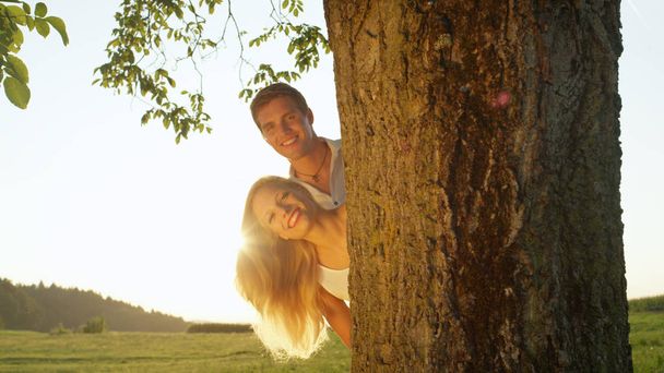 Yakın Lens Flare Up: bir yaz akşamı bir ağacın arkasından bakmak çift gülümsüyor. Genç erkek ve kadın bir ağaç gövdesi saklanıyor geçmiş parlayan yapraklara. Oğlan ve kız altın gün batımında romantik tarihte - Fotoğraf, Görsel