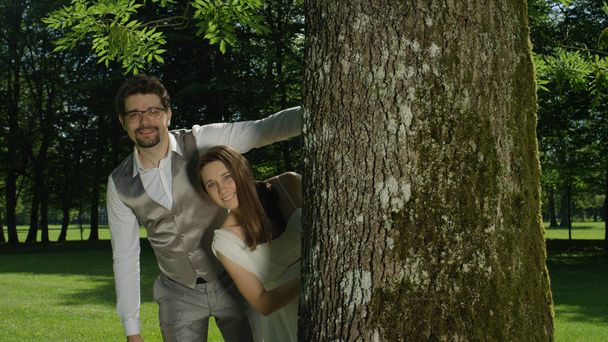 Hombre y mujer enamorados posando junto a un tronco de árbol en medio de un impresionante parque verde. Recién casados disfrutando de su sesión de fotos al aire libre después de una boda romántica. Pareja joven divirtiéndose en un jardín soleado
. - Foto, imagen