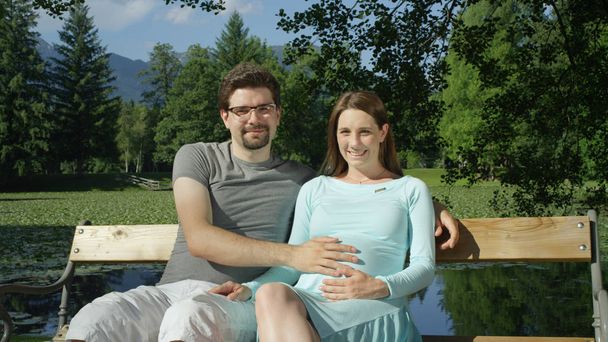 Portret atrakcyjna Młoda para siedzi na ławce w hipnotyzujący zielony Park w słoneczny dzień uśmiechnięty w aparacie. Rodzice mogą zrelaksować się na świeżym powietrzu w letni dzień. Mąż i żona zaczynają rodzinę - Zdjęcie, obraz