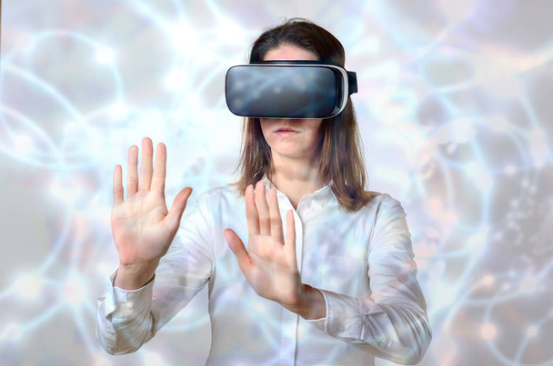 Mujer de camisa blanca tocando objetos invisibles en realidad virtual con sus dos manos, de pie en gafas 3D negras sobre fondo abstracto brillante. Retrato frontal de media longitud
 - Foto, imagen