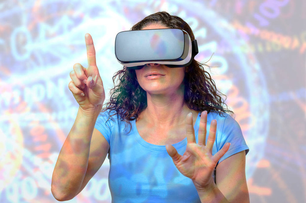 Nainen sinisessä t-paidassa käyttäen VR-liitäntää 3D-suojalaseissa, osoittaen etusormella näkymätöntä nappia. Edessä puolipitkä muotokuva vastaan abstrakti digitaalinen tausta projektio seinälle
 - Valokuva, kuva