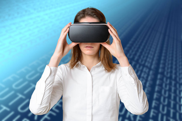 Jovem mulher de camisa branca ajustando óculos 3D na cabeça, segurando o dispositivo preto com as duas mãos, experimentando a realidade virtual. Retrato frontal de meio comprimento contra fundo digital abstrato azul
 - Foto, Imagem