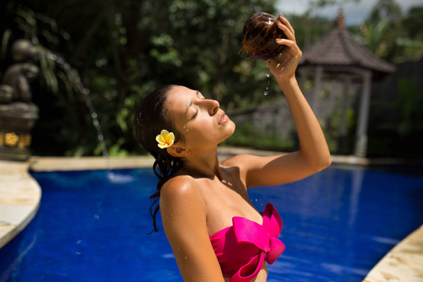 Sexy bruna sottile giovane donna che si annaffia con latte di cocco fresco in piscina con acqua cristallina. Royal resort tropicale relax
. - Foto, immagini