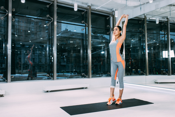 szczupły sprawny Azjatki stojąc na fitness Mata i rozciąganie w nowoczesnej siłowni - Zdjęcie, obraz