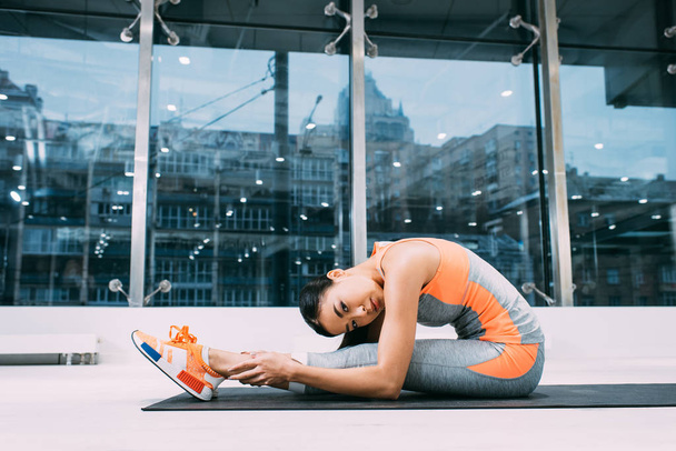 delgado asiático sportswoman haciendo estiramiento ejercicio en fitness mat y mirando cámara en deportes centro
 - Foto, imagen