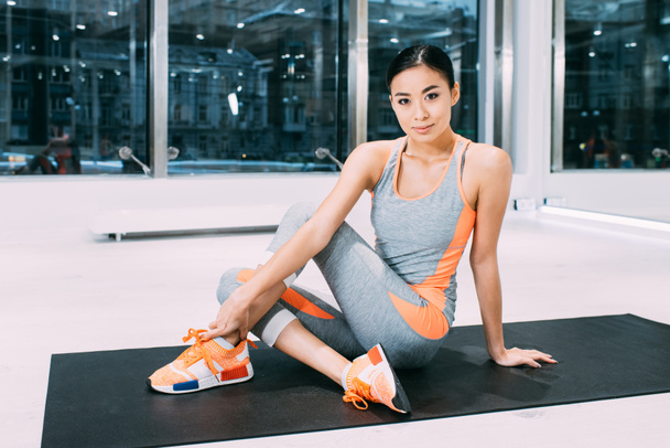 smiling asian girl sitting on fitness mat and looking at camera in modern sports center - Zdjęcie, obraz