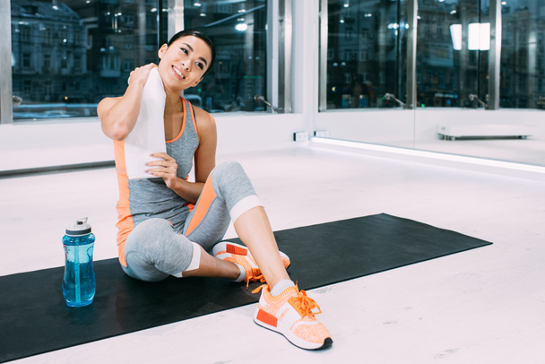 sonriente asiático chica sentado en fitness mat con toalla en moderno gimnasio
 - Foto, imagen