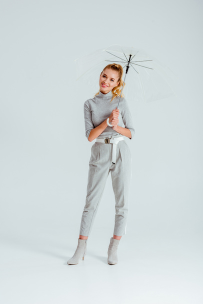 beautiful smiling woman in grey clothes posing with transparent umbrella on grey background - Foto, afbeelding