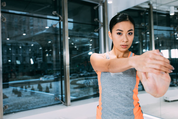 крупним планом вид на молоду азіатську дівчину, що стоїть, розтягує руку і дивиться на камеру в спортзалі
 - Фото, зображення