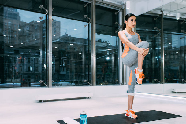 стройная азиатская девушка, стоящая на коврике для фитнеса и растягивающая ноги в современном спортзале
 - Фото, изображение