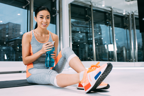 smiling asian girl sitting on fitness mat, holding sports bottle with water and looking at camera at gym - Fotografie, Obrázek