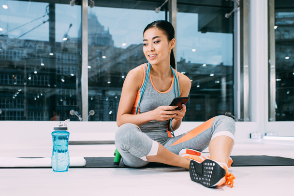 hymyilevä aasialainen urheilija istuu kunto matto ja tilalla älypuhelin urheilukeskuksessa
 - Valokuva, kuva