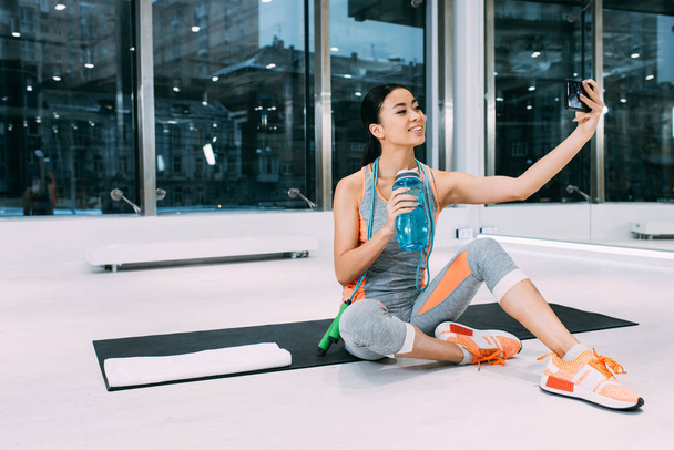 smiling asian girl sitting on fitness mat, holding sports bottle with water and taking selfie at gym - 写真・画像