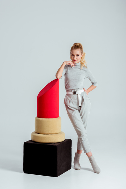 woman touching chin, posing near big red lipstick model and looking at camera on grey background - Foto, afbeelding
