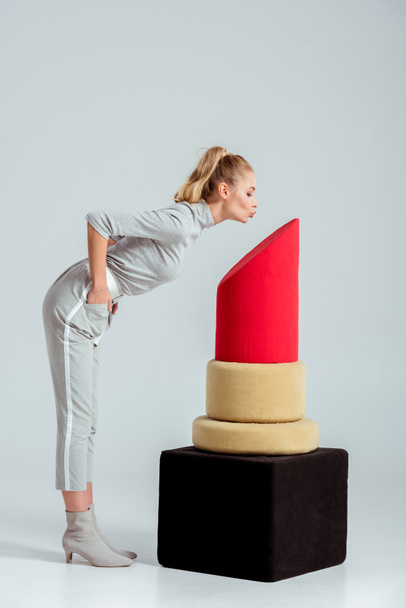Женщина целует воздух рядом с моделью красной помады на сером фоне
 - Фото, изображение