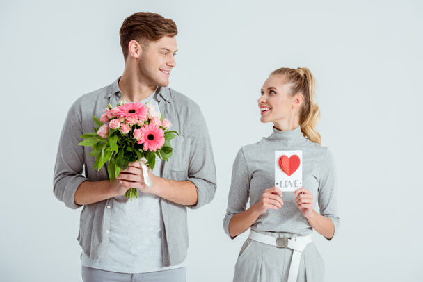 happy couple looking at each other and holding Valentines card and flower bouquet isolated on grey - Photo, Image