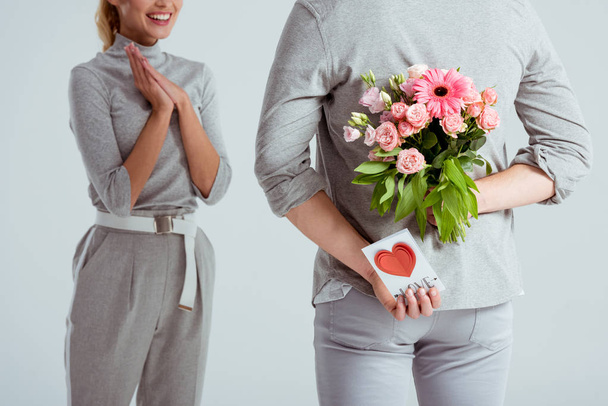 abgeschnittene Ansicht einer Frau mit gefalteten Händen, die auf einen Mann blickt, der Blumenstrauß und Valentinskarte hinter dem Rücken versteckt, isoliert auf grau - Foto, Bild