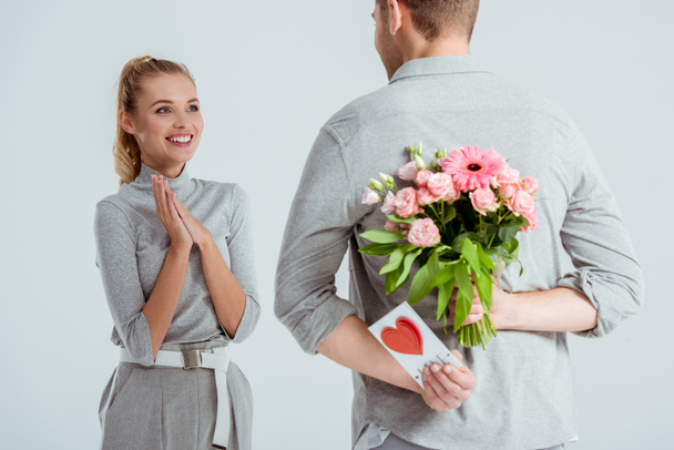 Женщина со сложенными руками, смотрящая на мужчину, прячущего букет цветов, и поздравительная открытка за спиной, изолированная на сером
 - Фото, изображение