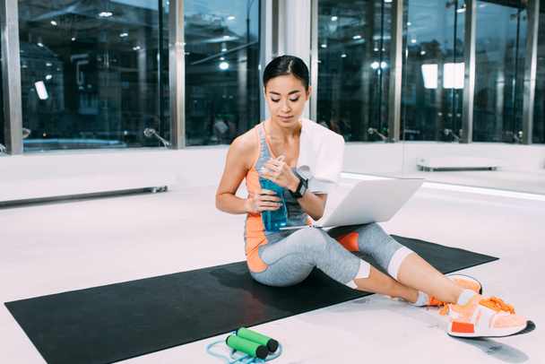 asian sportswoman sitting with laptop on fitness mat and holding sports bottle at gym - 写真・画像