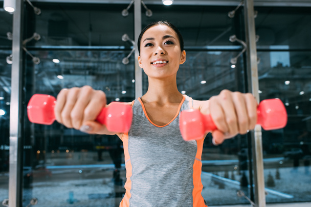 primer plano vista de atractivo asiático sportswoman sonriendo y haciendo ejercicio con dumbbells en gimnasio
 - Foto, Imagen