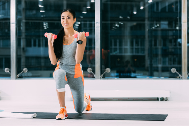 улыбающаяся азиатская спортсменка делает упражнения с гантелями на фитнес-коврике в спортзале
 - Фото, изображение