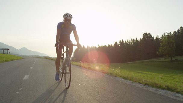 Молодий спортсмен їде на велосипеді через мальовничу літню природу на заході сонця. Красиві золоті сонячні промені освітлюють атлетичного чоловіка, розважаючись педалі свій велосипед уздовж асфальтової дороги
 - Фото, зображення
