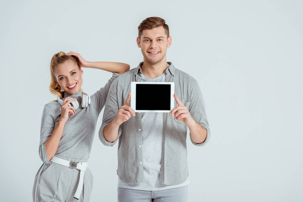 beautiful woman in headphones standing near man presenting digital tablet with blank screen isolated on grey - Photo, Image
