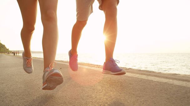 LOW ANGLE, LENS FLARE: Unknown man and woman jog in the sunny summer near blue ocean. Unrecognizable couple runs down sunny asphalt road leading past stunning coast. Summer holiday outdoor activity. - 写真・画像