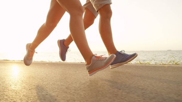 LOW ANGLE, LENS FLARE: Unrecognizable young couple running together near peaceful ocean on a beautiful summer afternoon. Active young people enjoy the sunny summer at the seaside and jog along coast. - Zdjęcie, obraz