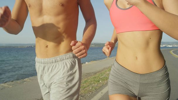 Close-up: Onherkenbaar jonge Kaukasische vrouw en man jog zonnige kust weg. Shirtless man en vrouw draagt Sport kleding uitvoeren op zomervakantie. Jonge joggers zonovergoten oceaan weg rennen - Foto, afbeelding