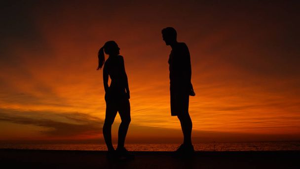 Alacsony szög, Silhouette: Vidám barátja és barátnője beszélt a strandon narancsszín napkelte. Imádnivaló fiatal házaspár mosolyog, míg a romantikus dátum a békés tengerparti lenyűgöző nyári este. - Fotó, kép