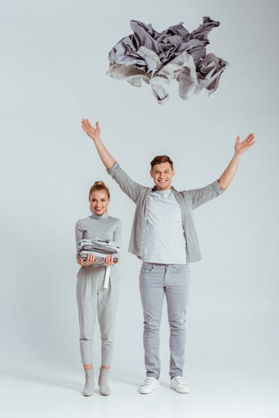 woman in grey outfit looking at camera and standing near man throwing pile of clothes in air on grey background - Photo, Image