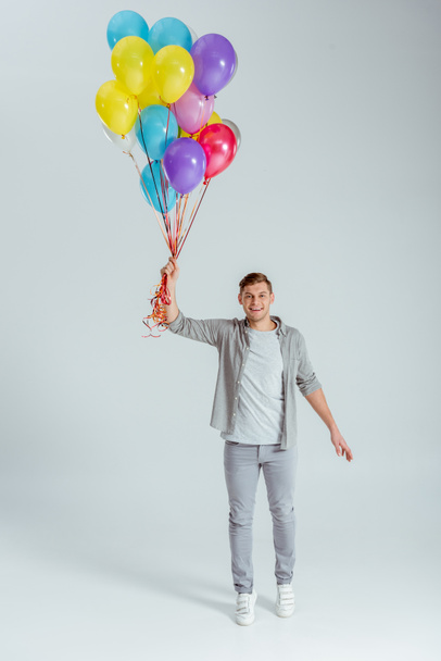 happy man holding bundle of colorful balloons and looking at camera on grey background - Фото, изображение