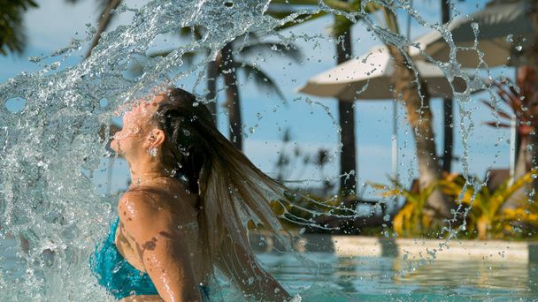 Close-up: Kaukasische meisje speels draait haar haren achteruit in zonnige exotische zwembad. Vrolijke jonge vrouw zwepen haar haar rug en sprays glinsterend druppeltjes van water in tropische zwembad op een warme zomerdag. - Foto, afbeelding