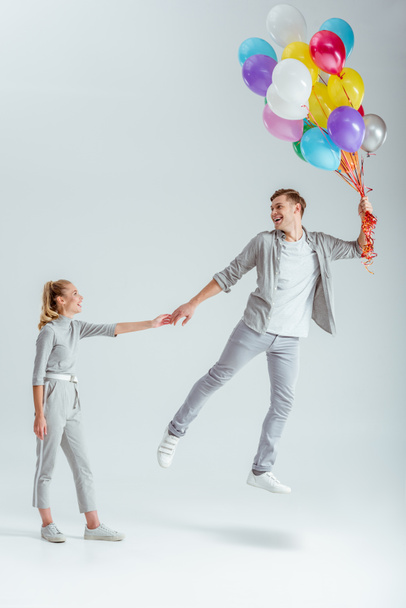 woman in grey clothing holding hand of happy man jumping in air with bundle of colorful balloons on grey background - Photo, Image