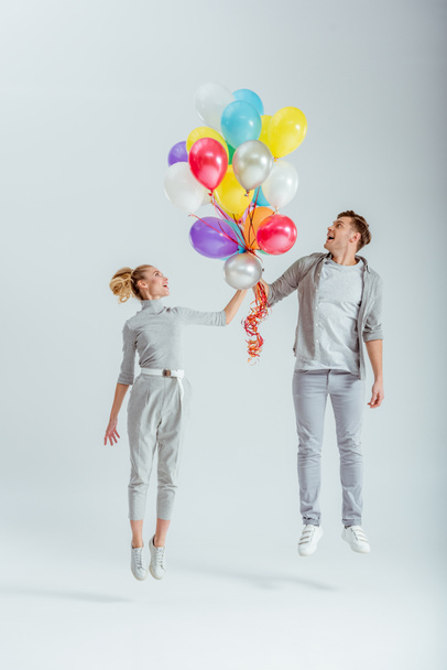 pareja excitada en ropa gris saltando en el aire con un paquete de globos de colores sobre fondo gris
 - Foto, Imagen